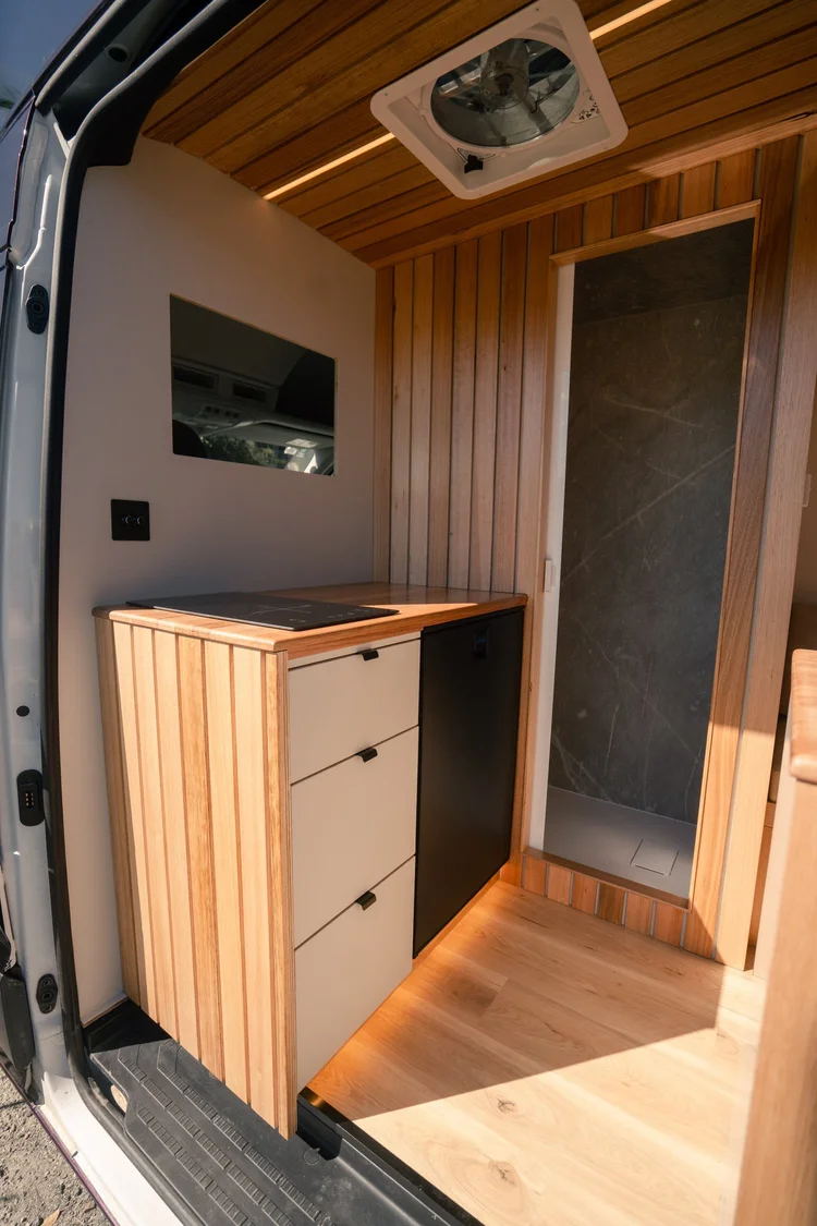 Galley with twin induction cooktop, timber benchtop and 95L fridge beside the shower door