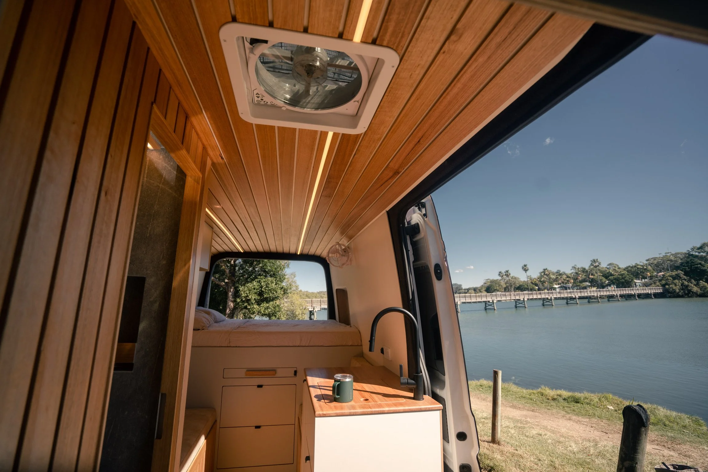 Morning light inside the Bondi Van Co campervan, a coffee cup on the timber bench and a jetty stretching across still water