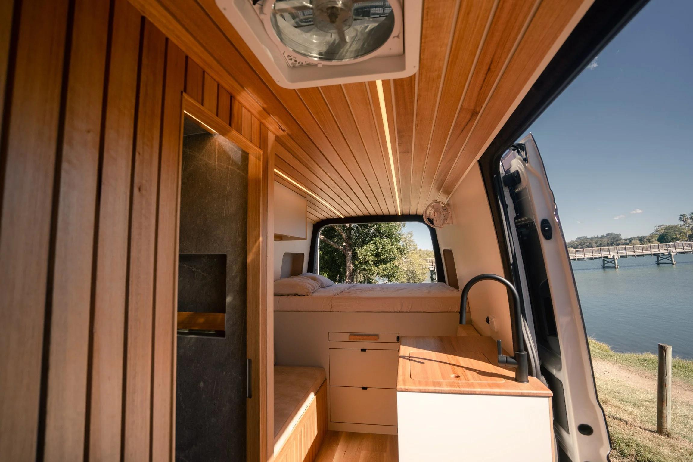 Interior of the campervan with timber-lined ceiling, stone shower door, bed and galley, side door open onto water